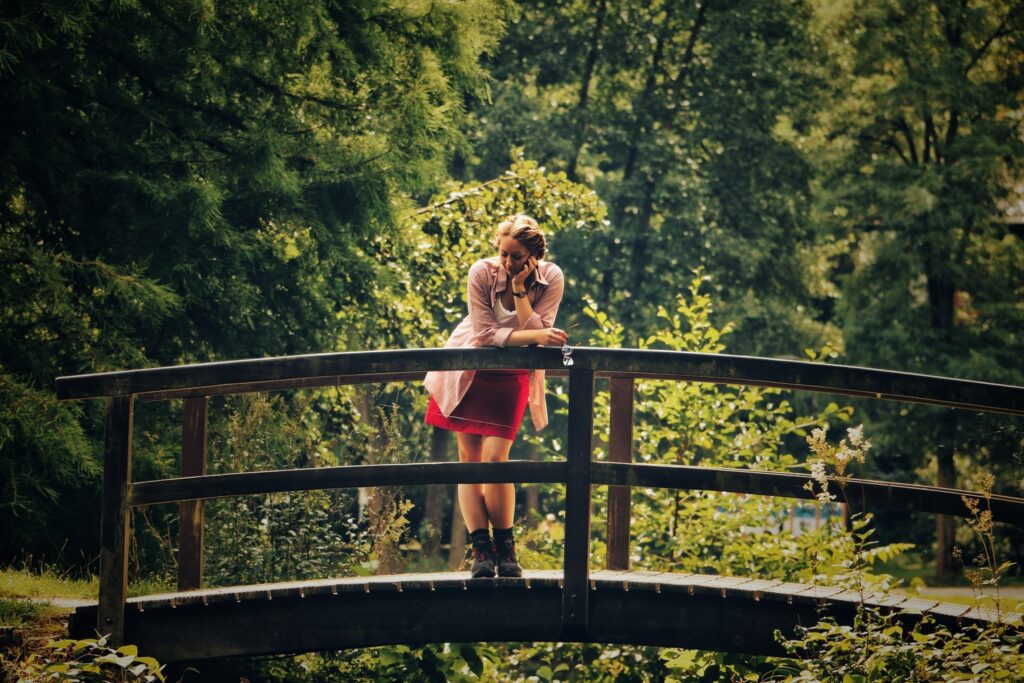 Girl standing on wooden arched bridge in a park, with green trees in the background.
